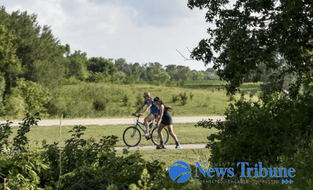 Trail in a green area with trees. Include two white women, one on a bike and the other one walking. News Tribune logo in blue